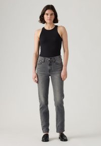 Črna rebrasta majica brez rokavov v kombinaciji z visokim pasom, izbledelimi sivimi džinsami in črnimi loaferji; model stoji pred nevtralnim ozadjem.