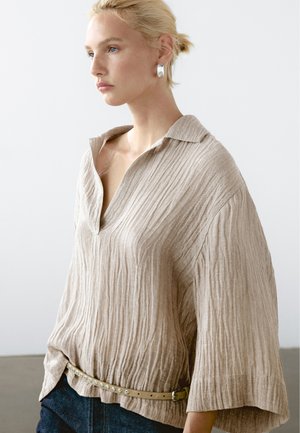 Top beige con texture, con scollo a V e colletto aperto, caratterizzato da maniche larghe e vita con cintura. Il modello indossa orecchini argentati oversized.