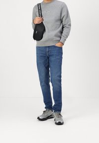 Grauer Pullover mit Logo, blaue Jeans und graue Sportschuhe. Hält eine schwarze Tasche mit weißen Akzenten. Leger outfit, verschiedene Texturen.