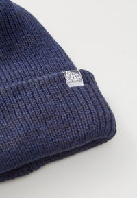 Caciula tricotată de culoare albastră închisă, cu textură în relief. Prezintă o etichetă mică pe pliul superior, care arată un logo și "Last Resort AB."