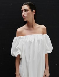 Γυναίκα με σκούρα μαλλιά χτενισμένα προς τα πίσω, φορώντας λευκό φόρεμα με αποκαλυπτούς τους ώμους και φουσκωτά μανίκια, σε μαύρο φόντο.