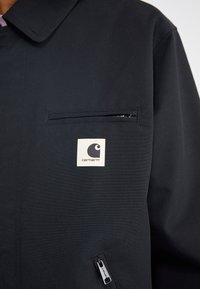 Zwarte jas gemaakt van duurzaam stof, voorzien van een ritssluitingzak, Carhartt-logo patch en een klassiek kraagontwerp.