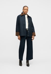 Γυναίκα με πλεγμένα μαλλιά, φοράει σκούρο μπλε σακάκι με καφέ γιακά και μανσέτες, λευκό τοπ, σκούρο τζιν με φαρδιές παντελόνες και μαύρα μποτάκια μέχρι τον αστράγαλο.