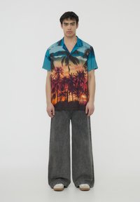 Camicia a maniche corte con una stampa colorata di tramonto con palme in tonalità blu e arancione, abbinata a pantaloni in denim grigi larghi e scarpe bianche.