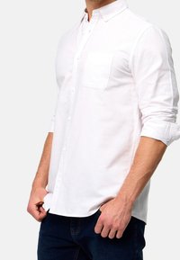 Chemise blanche à boutons, avec un col, une seule poche poitrine et des manches retroussées. Fabriquée en tissu léger ; texture lisse.