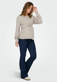 Bež pletena cardigan s kabelskim vzorcem, gumbi spredaj, oprijet pas z detajlom pentlje, valovit rob, v kombinaciji s temnimi džinsom in supergami.