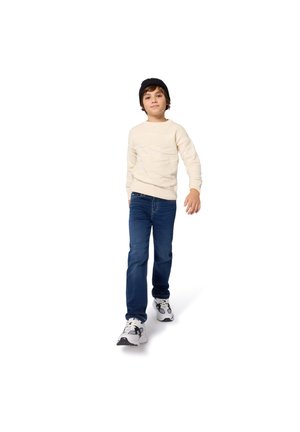 Suéter de punto crema con patrón texturizado, jeans de mezclilla azules y zapatillas negras. El niño camina contra un fondo blanco.