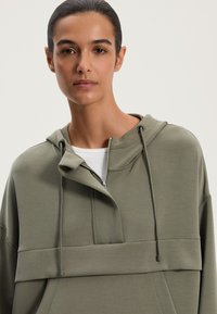 Olivovo zelená mikina s opaskom a nastaviteľnou šnúrkou. Má uvoľnený strih a hladkú, textúrovanú látku.