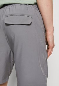 Šedé šortky vyrobené z lehkého materiálu s elastickým pasem a jednou zadní kapsou; hladký povrch a jemný lesk.