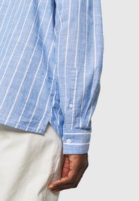 Camicia a maniche lunghe a righe blu chiaro realizzata in lino. Presenta strisce bianche, polsini con bottoni e un orlo curvo.