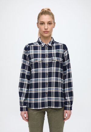 Une femme se tient de face, vêtue d’une chemise boutonnée à carreaux bleu marine, blanc et bleu, et d’un pantalon olive, sur un fond uni.
