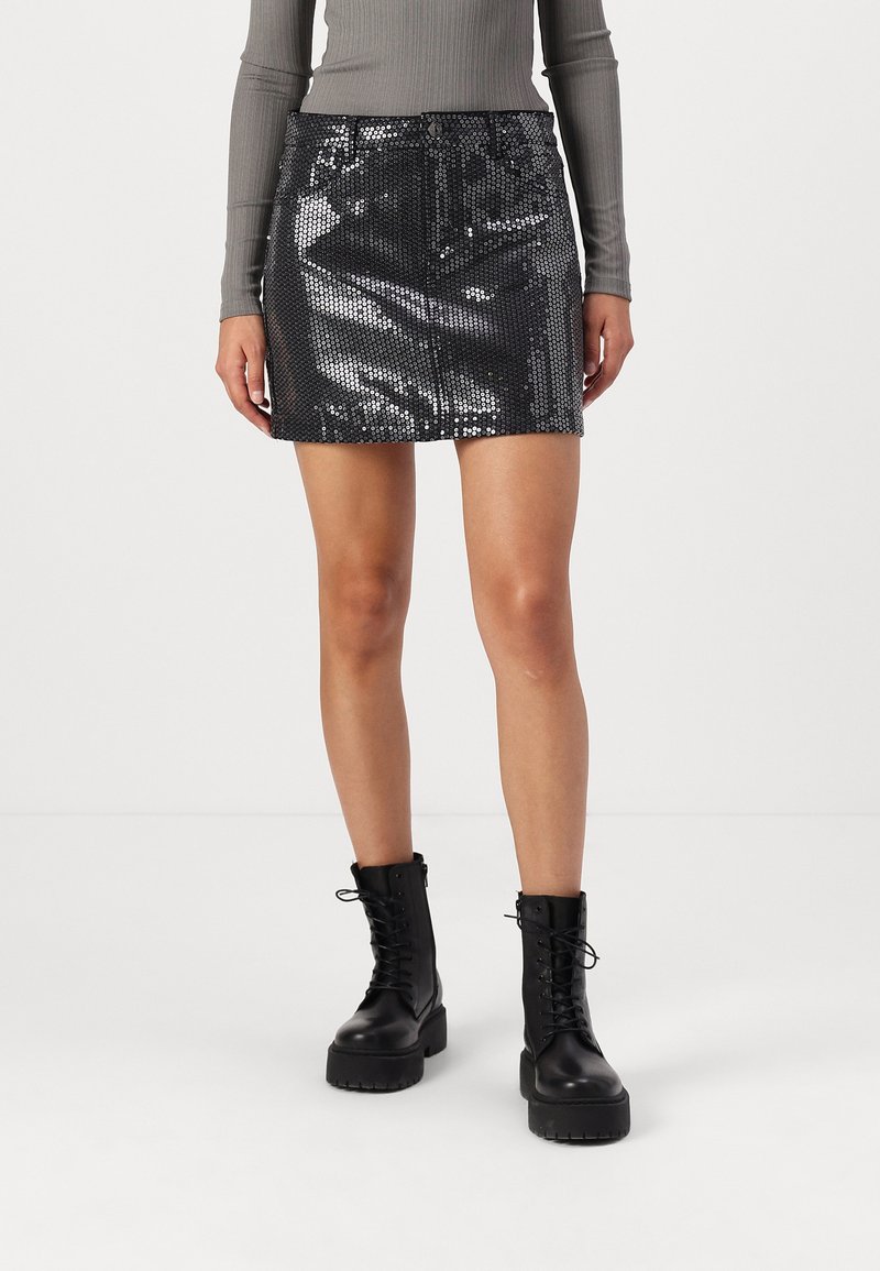 Zwarte glinsterende mini-rok met een aansluitend design en riemlussen, gecombineerd met hoge zwarte veterlaarzen. Glad, glanzend oppervlak.