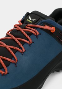 Μπλε σουέτ παπούτσι με μαύρες λεπτομέρειες, πορτοκαλί λαστιχένια κορδόνια ζικ ζακ και υφασμάτινη, αντιολισθητική σόλα. Εντυπωσιακό λογότυπο στη γλώσσα.