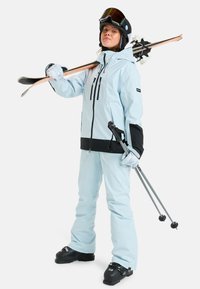 Jasnoniebieska kurtka narciarska z czarnymi akcentami, w połączeniu z pasującymi spodniami. Narty na ramieniu, kijki narciarskie w ręku, kask i gogle na głowie.