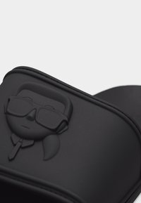 Μαύρο σανδάλι με φτέρνα με ανάγλυφο πρόσωπο που φοράει γυαλιά ηλίου, με αλογοουρά και γραβάτα στο λουράκι.