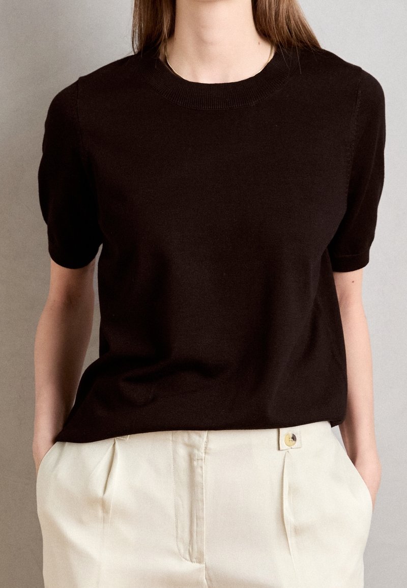 Žena ve černém krátkém svetr s krátkým rukávem zasunutém do krémových kalhot s vysokým pasem, s rukama v kapsách na jednobarevném pozadí.