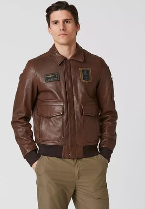 Uomo che indossa una giacca bomber in pelle marrone con toppe, in piedi con le mani nelle tasche, vestito con pantaloni khaki su uno sfondo semplice.