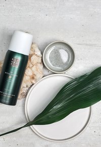 Flacone spray in alluminio verde con tappo bianco, piatto in ceramica grigia, base in pietra testurizzata, grande foglia verde e un piccolo piatto di liquido.