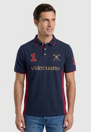 Hombre que lleva una camiseta polo azul marino con paneles laterales rojos, número 1 en las mangas y el pecho, y texto dorado "valecuatro POLO EDITION".