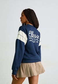 Sweat-shirt bleu marine avec des rayures blanches sur les manches et un logo. Présente des poignets et un ourlet côtelés. Assortie avec une jupe plissée beige.