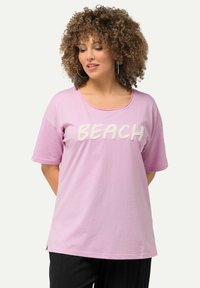 Lyserød t-shirt med korte ærmer med teksten "BEACH" i hvidt tekstureret bogstaver, rund halsudskæring og sideslids for bedre bevægelsesfrihed.