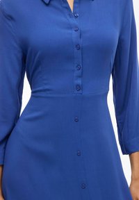 Modrá košela sukňa vyrobená z hladkej tkaniny; má golier, dlhé rukávy a priliehavý pás s rovnomerne rozmiestnenými gombíkmi.