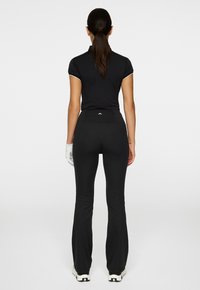Donna con ponytail scura, che indossa un top corto nero, pantaloni neri svasati, un guanto bianco sulla mano sinistra e scarpe bianche, di spalle.