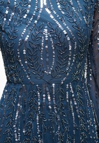 Marinblå klänning med intrikata silverpaljetter i bladmönster, slät tygtextur, figursydd midja och långa ärmar, som visar detaljerade dekorationer.