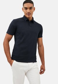 Marineblaues Poloshirt aus Baumwollstoff mit klassischem Kragen, kurzen Ärmeln und einem kleinen gestickten Logo auf der Brust. Kombiniert mit weißen Hosen.