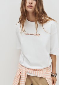 Weißes Baumwoll-T-Shirt mit Rundhalsausschnitt und kurzen Ärmeln; der Druck auf der Vorderseite lautet "ICH BRAUCHE EINE EISCREME" in Braun. Gestreifter oranger Stoff um die Taille gebunden.