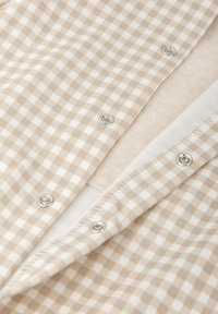 Tejido de cuadros beige y blanco con broches metálicos, superpuesto sobre suave forro polar beige y forro blanco, que muestra una textura suave y una costura ordenada.