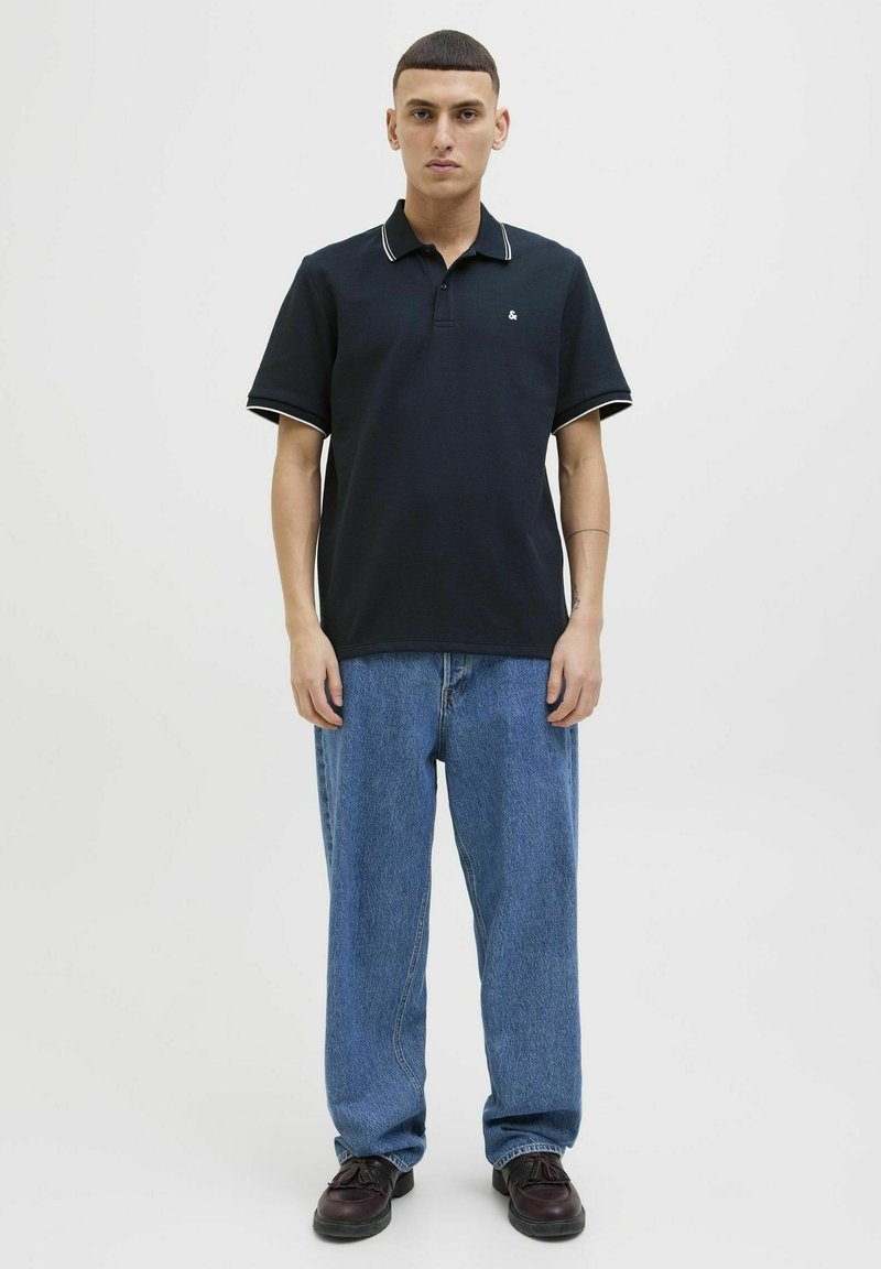 Polo shirt bleu marine avec col et poignets à rayures blanches, associé à un jean en denim bleu clair ample et des chaussures foncées. Texture lisse, logo minimal.