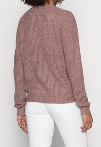 Person som bär en lila stickad tröja med långa ärmar och vita tajta jeans, sedd bakifrån mot en enfärgad bakgrund.