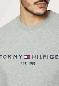 Sudadera gris con cuello redondo, con texto bordado "TOMMY HILFIGER EST. 1985" y un pequeño logo de colores en el centro.