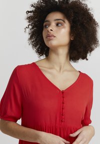 Robe rouge à manches courtes, décolleté en V, avec des boutons le long du devant. Fabriquée dans un tissu léger avec une finition texturée.