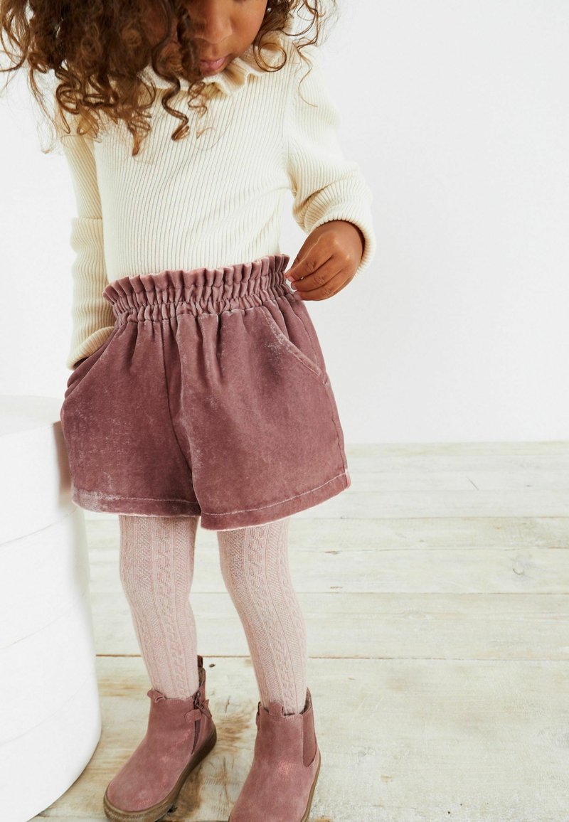 Shorts en velours rose avec une ceinture élastique et des poches latérales, assortis à des collants roses à motifs et des bottines roses assorties.