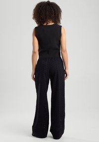 Mustat ilmanhihoiset neulepaidat yhdistettynä leveälahkeisiin mustiin housuihin, joissa on tekstuuri ja rento istuvuus, nähtynä takaa.