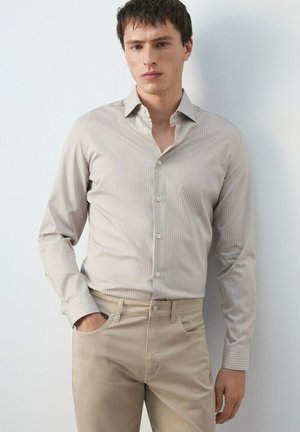 Hombre con pantalones ajustados beige y camisa de botones a rayas beige claro, de pie contra una pared blanca con una mano en el bolsillo.