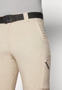 Pantalones cargo beige con un bolsillo lateral con cremallera, tela ligera con una textura suave y un cinturón negro ajustable.