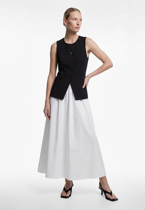 Kobieta ubrana w czarną kamizelkę bez rękawów, zapinaną na guziki, długą białą spódnicę i czarne sandały na obcasie, stojąca z jedną ręką na biodrze.