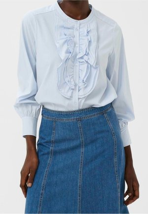 Femme portant un chemisier bleu clair à manches longues avec un devant à volants et une jupe en jean bleue taille haute avec des coutures verticales.