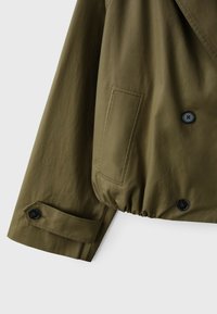 Manche de veste vert olive avec poignet boutonné, poche avant et deux boutons noirs sur la partie froncée de l'ourlet.