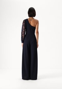Navy jumpsuit with one bare shoulder and one long lace sleeve, featuring a tailored fit and wide-leg design. Back zip closure visible.