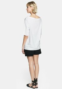 Biała, oversize'owa koszulka z szerokim dekoltem, zestawiona z czarnymi szortami i czarnymi płaskimi butami na sznurówki, widok od tyłu. Gładki, casualowy materiał.