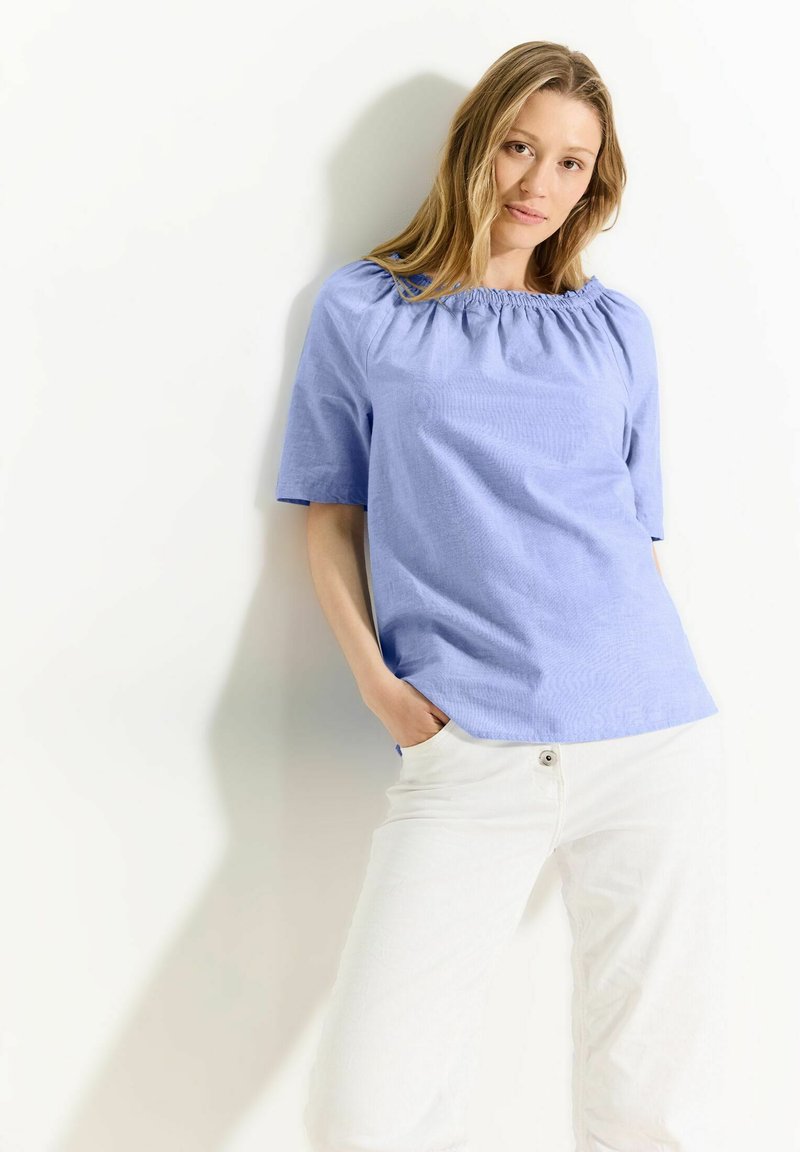 Himmelbluse mit kurzen Ärmeln und gerafftem Halsausschnitt, kombiniert mit weißen Hosen. Der Stoff wirkt glatt und leicht.
