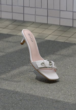 Sandalia mule de tacón blanco con punta cuadrada, detalle de hebilla plateada y nombre de la marca "RAID" visible en la plantilla, colocada sobre pavimento gris.