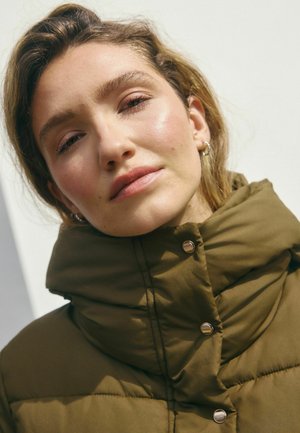 Jeune femme avec un maquillage léger portant une veste matelassée olive avec un col haut sur un fond clair.