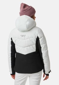 Giacca imbottita con parte superiore bianca e parte inferiore nera. Presenta un cappuccio, un design a chevron e un logo sul retro. Berretto a maglia di colore rosa.