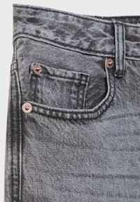 Gros plan sur la poche avant d'un jean denim gris avec une petite poche à monnaie, une couture orange, des rivets en métal et une passants de ceinture.