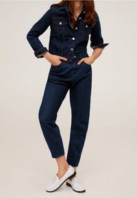 Chaqueta de mezclilla oscura con botones plateados y dos bolsillos en el pecho, combinada con unos jeans de tiro alto a juego y zapatillas blancas tipo slip-on.